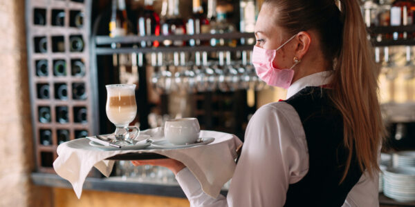 A female Waiter of European appearance in a medical mask serves Latte coffee.