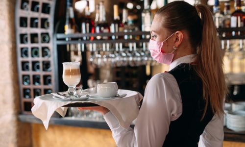 A female Waiter of European appearance in a medical mask serves Latte coffee